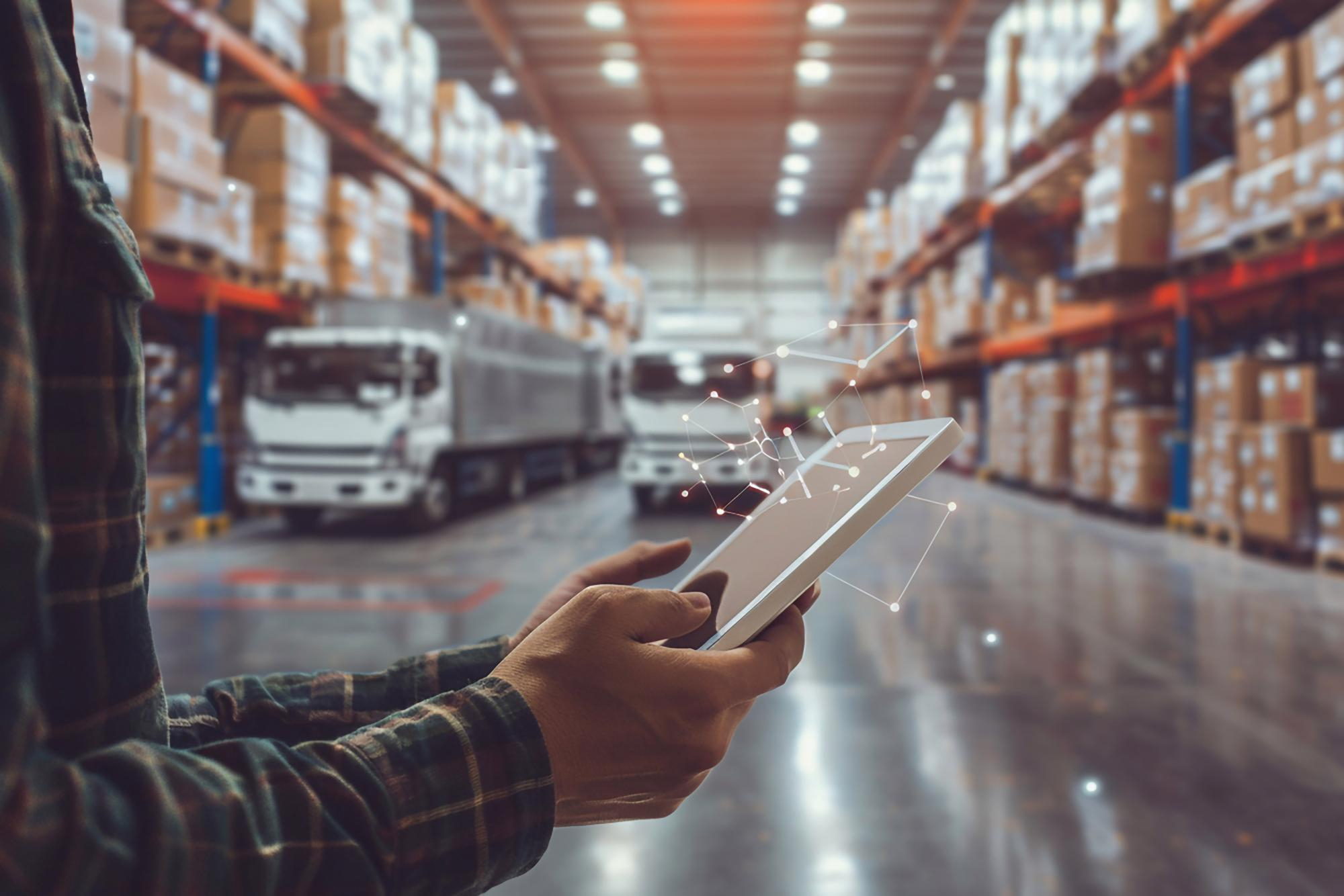 Planista logistyczny z tabletem w alejce magazynowej, ciężarówki w tle.
